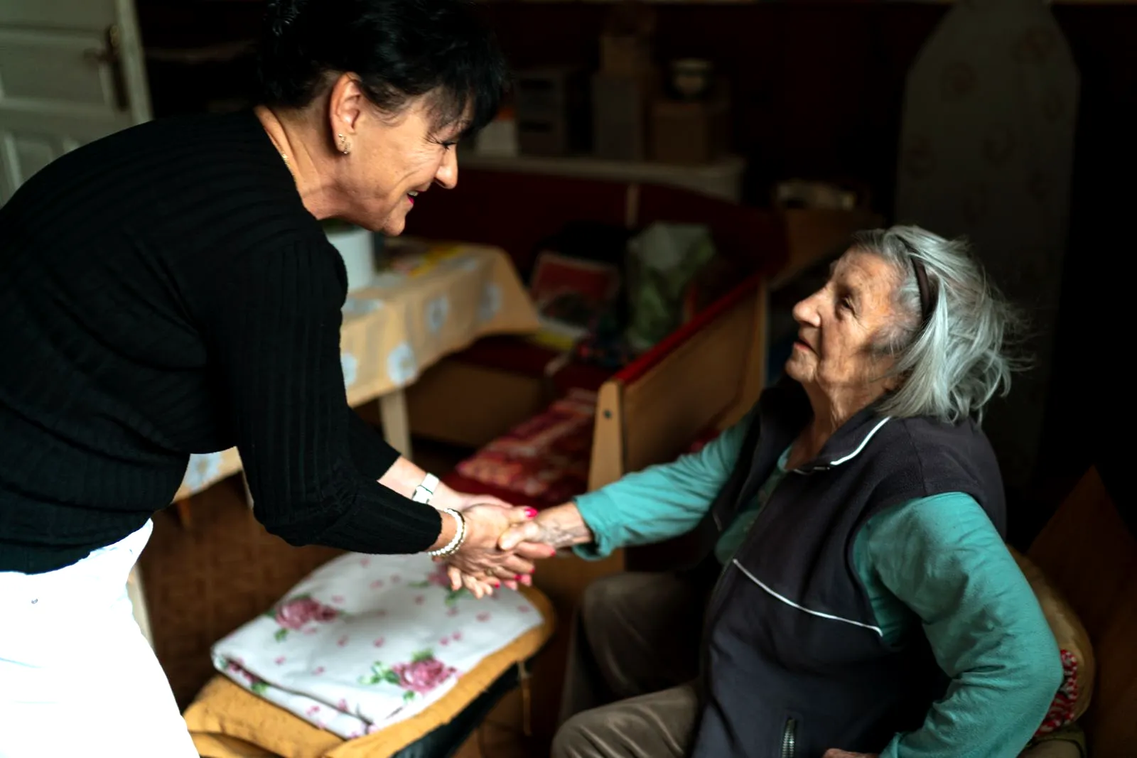 Eine Betreuungskraft in Arbeitskleidung begrüßt eine ältere Frau in deren Wohnung und reicht ihr die Hand.