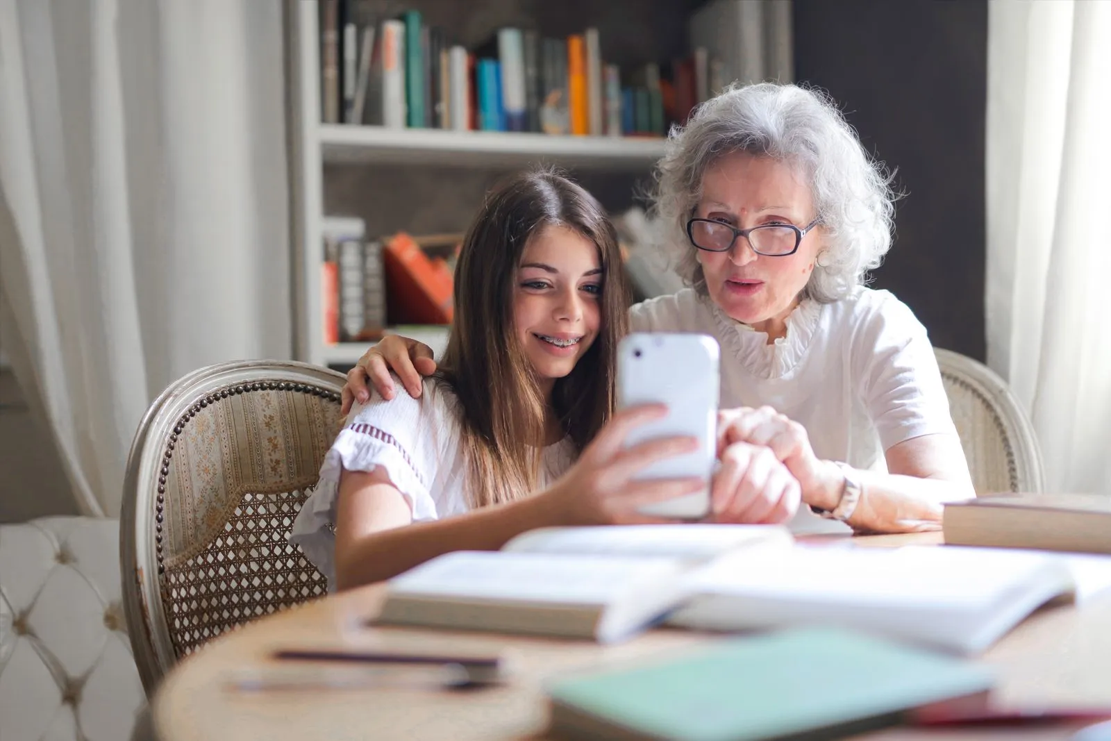 Eine ältere Frau und ihre Enkelin sitzen zu Hause am Tisch und schauen gemeinsam auf ein Smartphone.